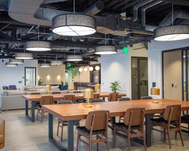 The image shows a modern office space featuring multiple tables, chairs, and seating areas under well-lit pendant lights.