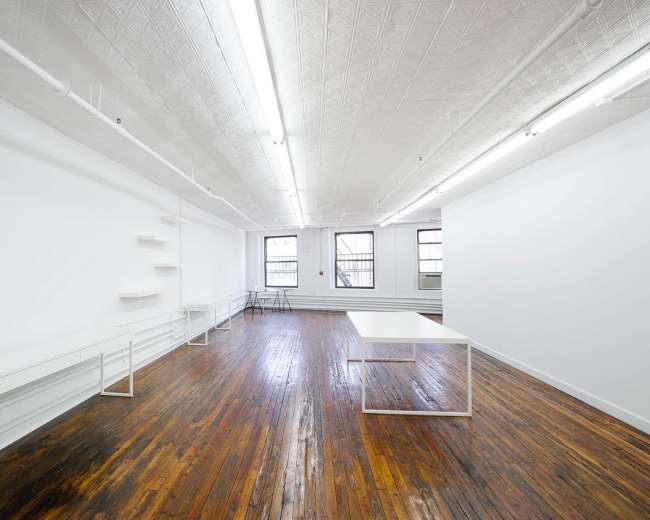 The image shows a spacious, empty room with wooden floors, white walls, and minimal furnishings, including a table and shelves.