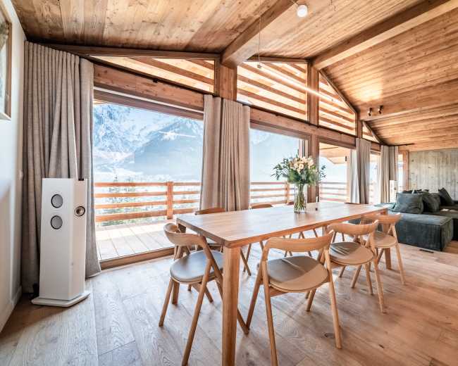 The image shows a wooden dining area with a table and chairs, large windows offering a view of mountains, and a cozy seating area adjacent to it.