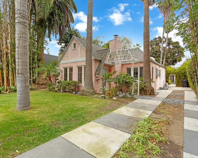 A pink bungalow is surrounded by lush greenery and palm trees, with a stone pathway leading to the entrance.
