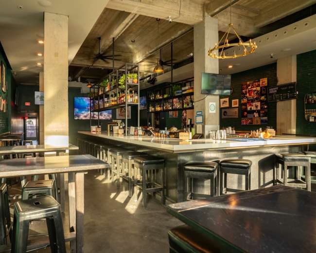 The image shows a modern bar interior with a long countertop, high stools, and shelves lined with bottles against a backdrop of green walls and large windows.