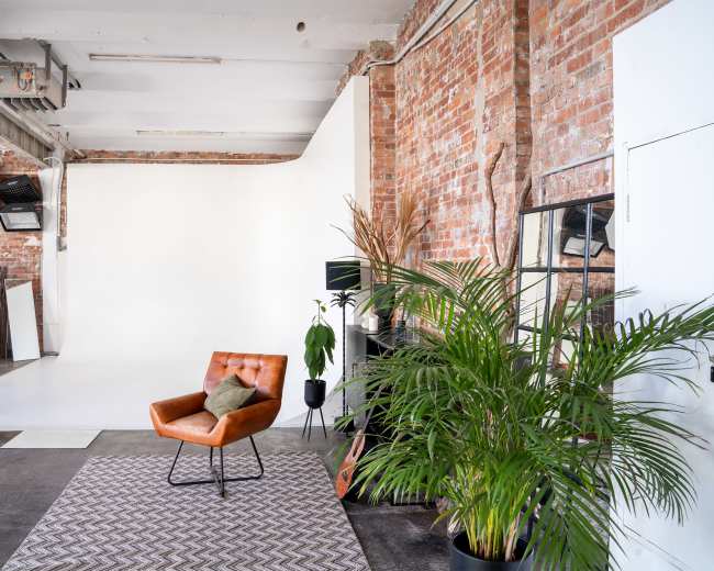 A minimalist photography studio featuring a brown leather chair on a patterned rug, a potted plant, and a backdrop of white and exposed brick walls.