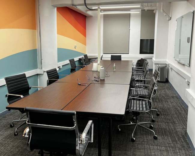 The image shows a modern conference room with a long table, ergonomic chairs, and colorful wall decor.