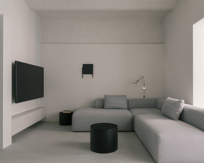 The image shows a minimalist living room with a gray sectional sofa, a black round coffee table, and a wall-mounted television.