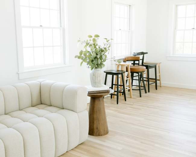 A sectional sofa is positioned near a set of tall stools and a small wooden side table, with a vase of greenery on top, in a bright, minimalist room.