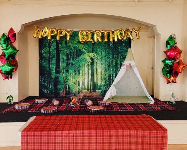A birthday celebration setup features a forest-themed backdrop, a small tent, decorated with balloons in green and red, and a checkered tablecloth.