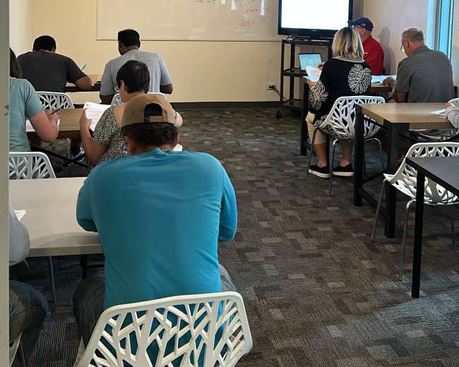 A group of people is seated at desks in a classroom, facing a whiteboard and a screen at the front of the room.