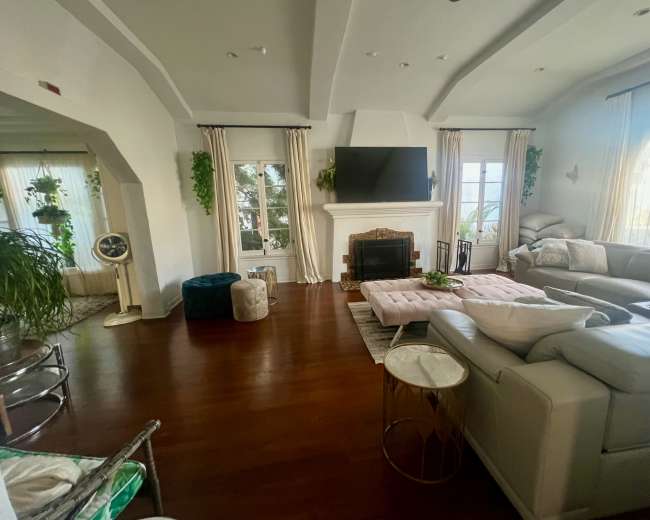 The image shows a spacious living room with a fireplace, large windows, and modern furniture arranged around a central seating area.