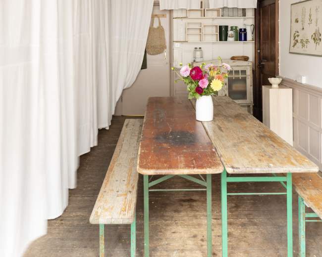 A long, rustic wooden table with benches sits in a bright, airy room adorned with white curtains and dried flowers hanging overhead.