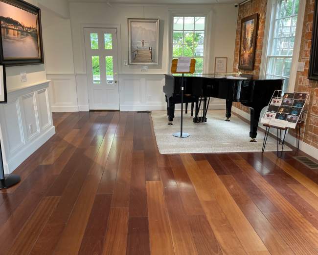 The image shows a room with wooden flooring, a black piano, framed wall art, windows, and a small area rug.