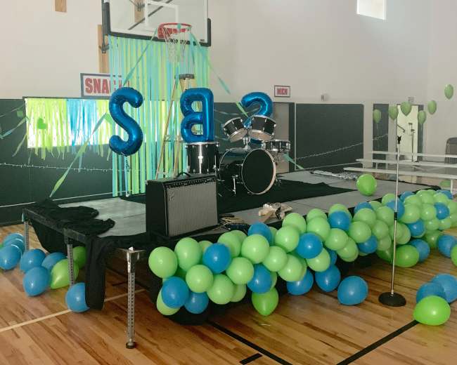 A decorated stage with a drum set and colorful balloons in shades of blue and green is set up in a gymnasium.