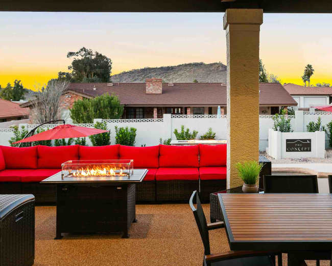 A patio area with a red seating arrangement, a fire pit in the center, a dining table, and views of a neighborhood and hills in the background.