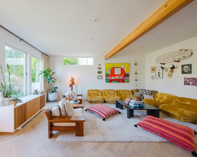 A spacious living room with large windows, a mix of seating options in yellow and neutral tones, and decorative artwork on the walls.
