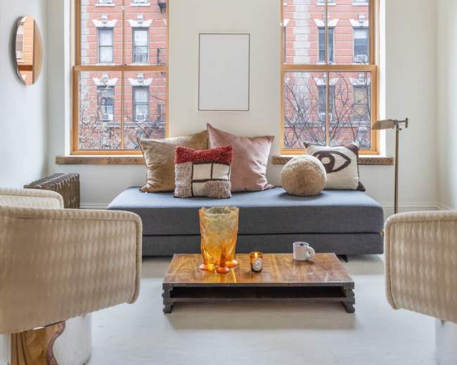 A cozy living room features a gray sofa with decorative pillows, a wooden coffee table, and large windows displaying a brick building outside.
