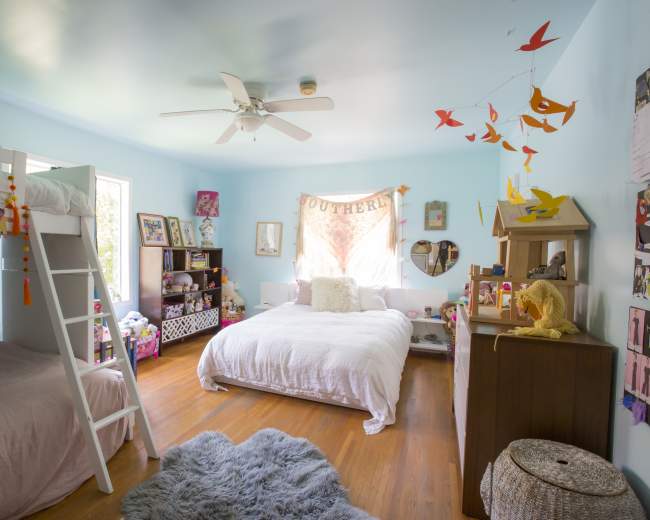 The room features a light blue wall, a bunk bed, a queen-sized bed, and decorated with colorful wall hangings and a mix of furniture.