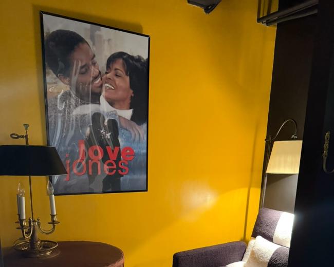 A cozy corner features a black and white upholstered chair beside a round table with a fringed edge, illuminated by a lamp, and a vibrant yellow wall adorned with a "Love Jones" movie poster.