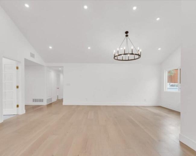 A spacious, empty room features light-colored wooden flooring, white walls, and a chandelier overhead.