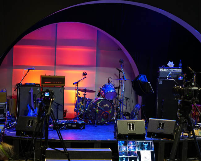A stage setup with musical instruments, amplifiers, and cameras arranged in a semi-circular backdrop.
