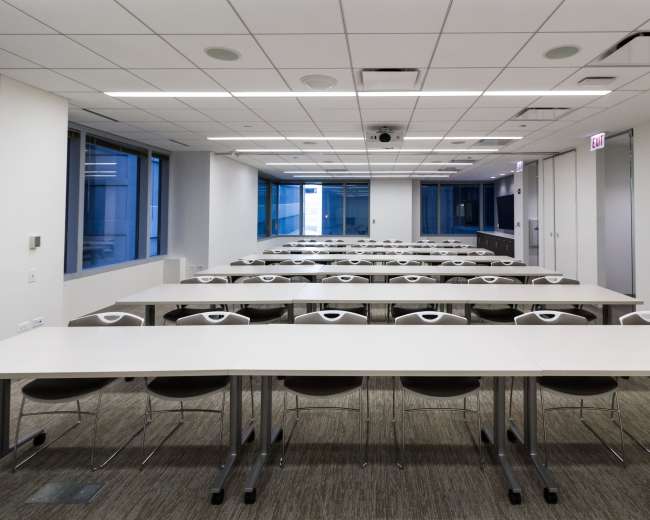 A spacious classroom features multiple rows of white tables and chairs, illuminated by natural light from large windows.