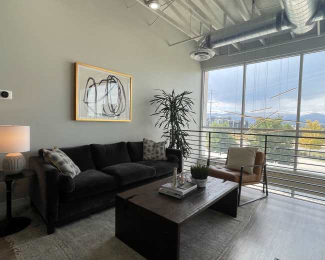 A modern living room with a black sofa, a wooden coffee table, a floor lamp, and large windows overlooking a scenic view.