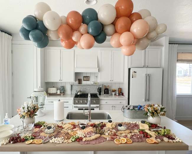 A beautifully arranged charcuterie board is displayed on a kitchen island, topped with a decorative balloon garland.