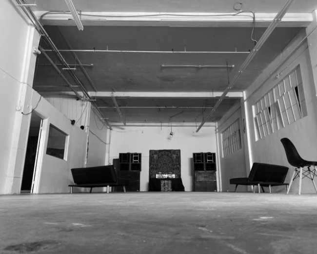 The image shows a spacious, empty room with two black sofas facing a central area that features a decorative piece and sound equipment.
