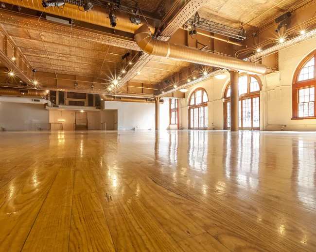 A spacious, empty room with polished wooden floors and large windows allowing natural light.