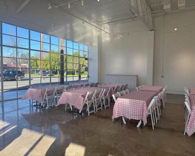 The image shows a spacious indoor venue with long tables covered in pink-and-white checkered tablecloths set up near large windows letting in natural light.
