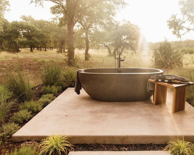 A modern freestanding bathtub sits on a concrete platform surrounded by greenery in a natural setting.