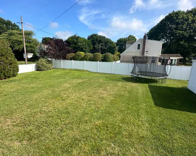 A spacious green backyard features a trampoline and a white fence, bordered by trees and houses in the background.