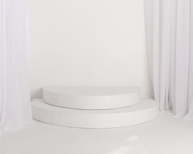 The image depicts a minimalist white stage with two circular platforms, surrounded by sheer white curtains.