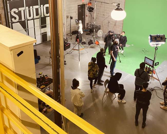 A film crew works in a studio setting with a green screen and various lighting equipment.