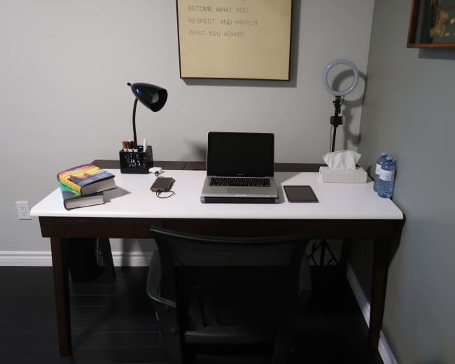 A minimalistic desk setup features a laptop, a smartphone, a tissue box, water bottle, books, and a lamp against a gray wall.