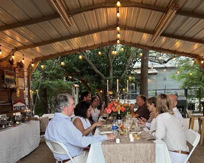 A group of people sits around a table decorated with flowers and candles in a covered outdoor dining area.