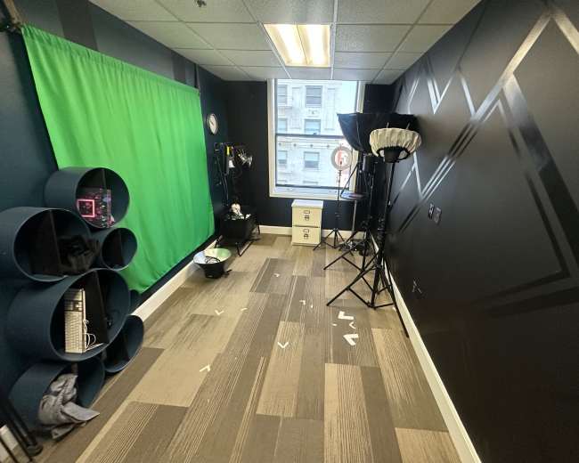 The image shows a small studio space with a green screen, lighting equipment, and a desk with storage drawers.