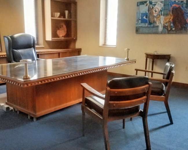 The image shows a simple office with a large wooden desk, two wooden chairs, and bookshelves against the walls.