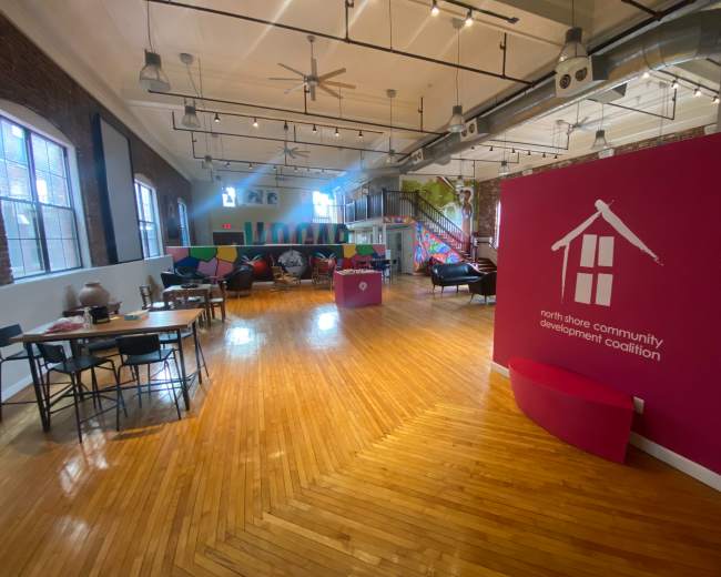 The image shows a spacious community center interior with wooden floors, colorful seating areas, and a prominent pink wall displaying the logo of the North Shore Community Development Coalition.