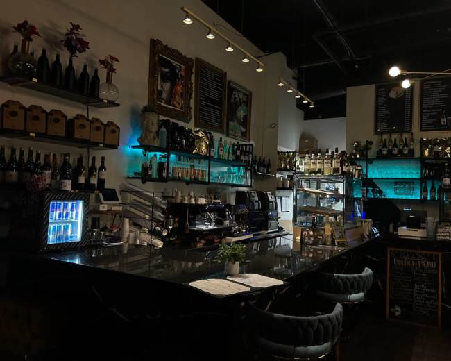 The image shows a dimly lit bar counter with shelves displaying various bottles and a coffee machine, accompanied by a menu board and decorative art on the walls.