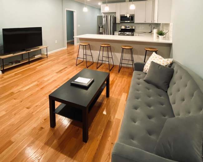 The image shows a modern living room and kitchen area featuring a gray couch, a black coffee table, bar stools at a kitchen island, and a large television on a stand.