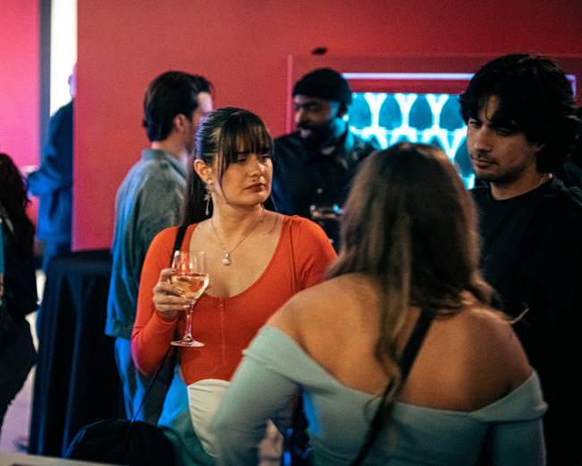 A crowded social event features a group of people mingling, with one woman holding a drink and wearing an orange top in the foreground.