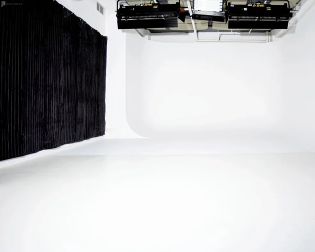 The image shows a photography studio with a white seamless backdrop, black curtains on one side, and studio lights mounted overhead.