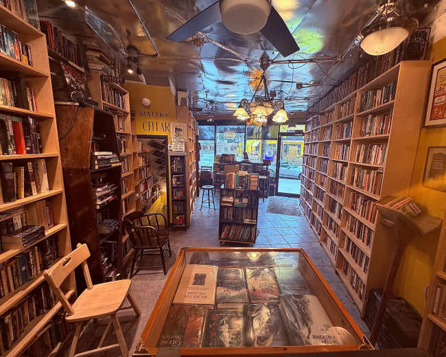 The image shows a cozy bookstore interior with shelves lined with books and a glass-topped display table in the foreground, while large windows allow natural light to illuminate the space.