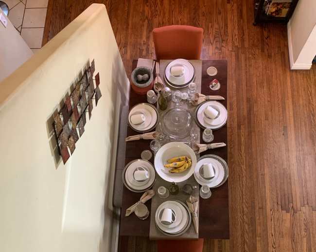 A dining table set for a meal, featuring plates, glasses, utensils, and a centerpiece bowl with bananas, surrounded by orange chairs.