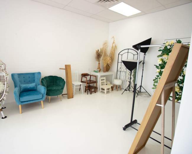 The image shows a bright studio space featuring a variety of chairs, decorative plants, and a podium, set against a white backdrop.
