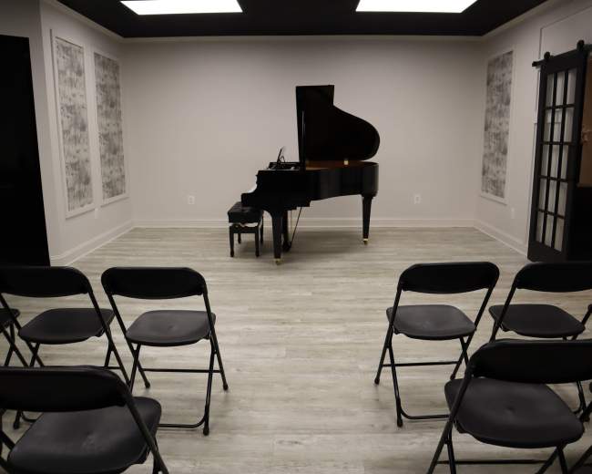 A grand piano stands in the center of a spacious room with black folding chairs arranged in rows facing it.