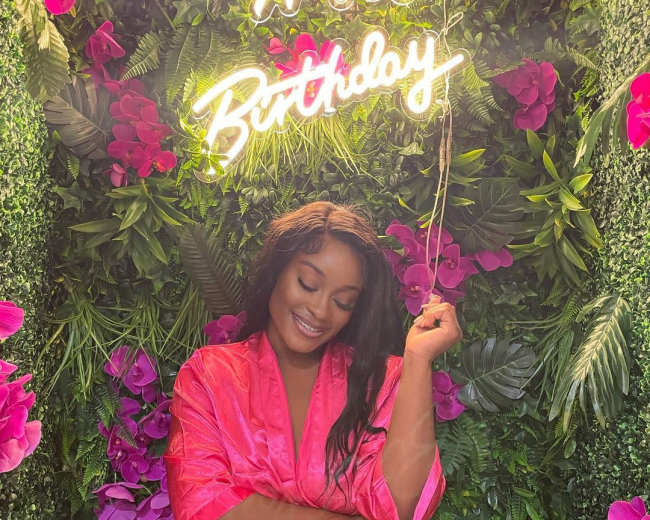 A woman in a pink robe stands in front of a greenery backdrop adorned with pink flowers and a neon sign that says "Happy Birthday."