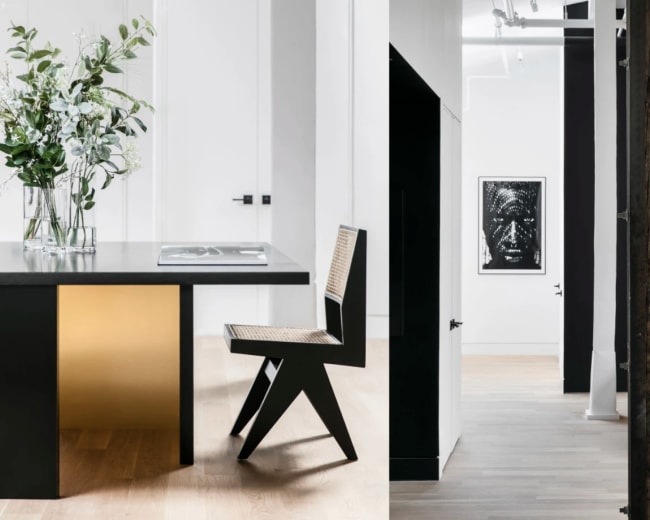 A split scene featuring a modern kitchen area with a sleek table and a chair, and a hallway leading to a room with a framed artwork and minimalist decor.