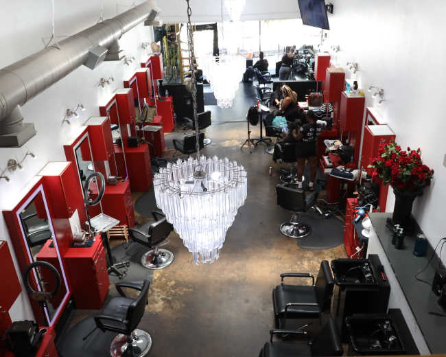 The image shows an interior view of a salon featuring red styling stations, black chairs, a large chandelier, and several stylists working on clients.