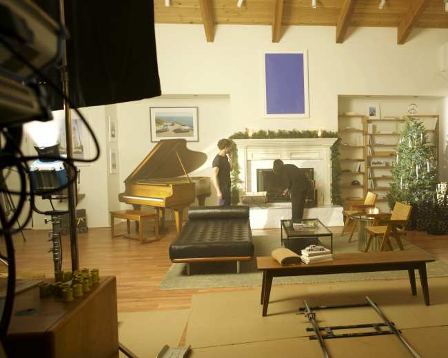 A living room scene features a grand piano, a decorated Christmas tree, and modern furniture, with two individuals engaged in activity near the fireplace.