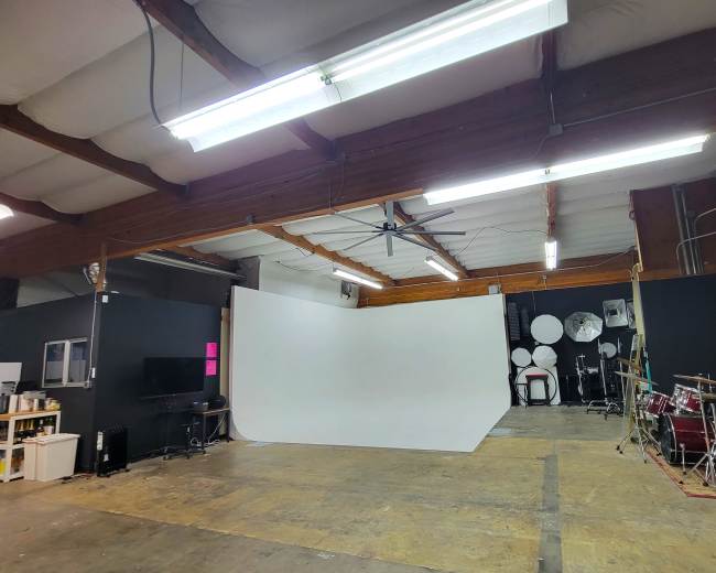 The image shows a spacious indoor studio with a white backdrop, some lighting fixtures, a television, and a drumming setup.